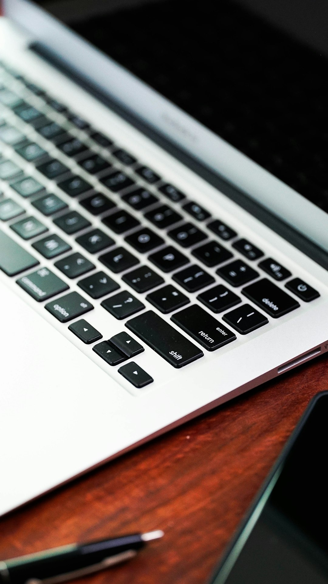 A cropped photo of a silver laptop on a wood-grained table top. It shows the bottom part of the screen, the right half of the keyboard, and part of the wrist rest. There's a black retractable ballpoint pen on the desk next to the computer.